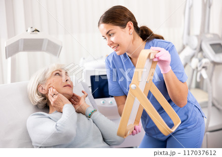 Young female dermatologist showing elderly female client result of facial beauty procedure in mirror in esthetic medicine center Young female dermatologist showing elderly female client result of facial beauty procedure in mirror in esthetic medicine center 117037612