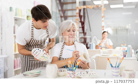 Guy assisting senior woman painting ceramic mug in pottery class Guy assisting senior woman painting ceramic mug in pottery class 117037755