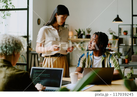 Young Black man and Asian woman wearing casual outfits having conversation while unrecognizable programmer coding on laptop in modern office 117039245