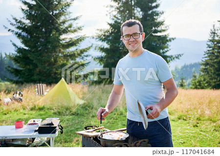 Man tourist stands outdoors, smiling while cooking at grill with pan and lid. Behind him, yellow tent and camping setup, surrounded by tall pine trees and scenic mountain landscape. 117041684