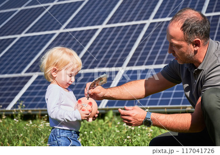 Modern father and his child putting cash into piggy bank to save money. Little kid interested in saving money for new toy. Man and his son putting money to piggy bank on background of solar battery. Modern father and his child putting cash into piggy bank to save money. Little kid interested in saving money for new toy. Man and his son putting money to piggy bank on background of solar battery. 117041726