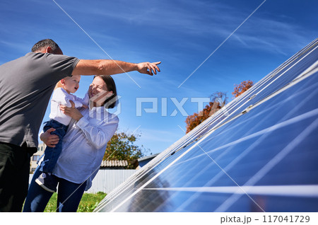 Father showing potential of alternative energy to his family. Young family looking at their investment for future. Side view of interested kid and his parents next to solar station. 117041729