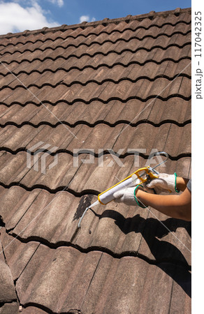 Worker man using silicone sealant adhesive  to fix crack of the old tile roof. 117042325