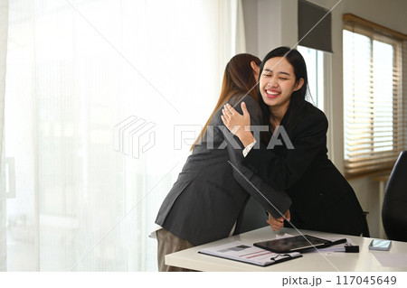 Happy cheerful businesswomen standing in office and embracing each other, celebrating good news 117045649