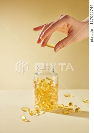 A hand holding a yellow vitamin pill in space, below is a transparent unlabeled vitamin jar with many pills scattered around. Yellow background photo was taken from front, designing space 117045744
