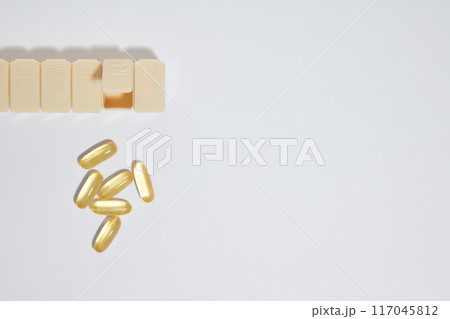 A yellow pill box placed in the top left corner, below is a small cluster of some yellow fish oil capsules, flat lay on the white countertop. Blank right side for designing and adding text 117045812