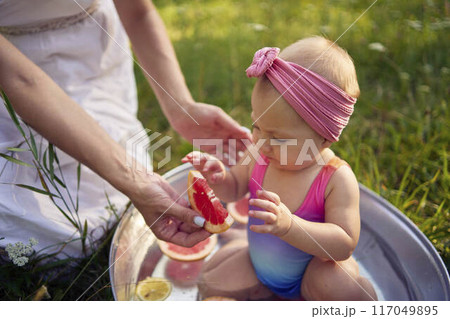 baby in metal bath in summer garden watching mother and teenage sister eat grapefruit 117049895