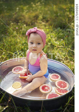 a baby in a metal bath in a summer garden plays with grapefruit and lemon slices a baby in a metal bath in a summer garden plays with grapefruit and lemon slices 117049904