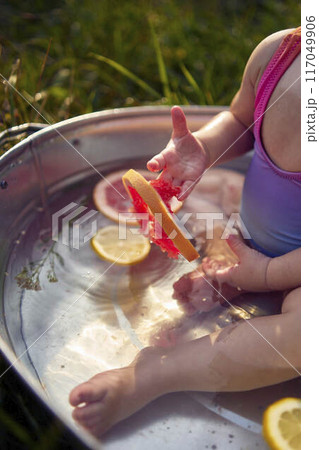 a baby in a metal bath in a summer garden plays with grapefruit and lemon slices 117049906