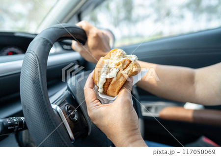 Asian lady holding hamburger to eat in car, dangerous and risk an accident. 117050069