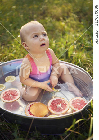 a baby in a metal bath in a summer garden plays with grapefruit and lemon slices 117050096
