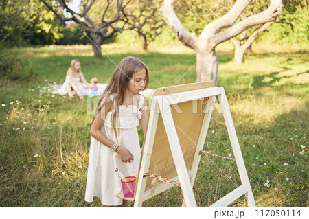 mother and baby sister watch as  pre-teen girl in a summer dress paints a picture in the garden on an easel 117050114