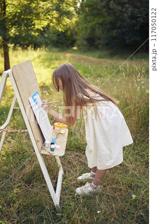 pre-teen girl in a summer dress paints a picture in the garden on an easel pre-teen girl in a summer dress paints a picture in the garden on an easel 117050122
