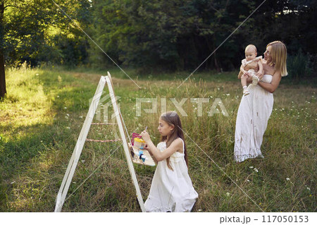 mother and baby sister watch as pre-teen girl in a summer dress paints a picture in the garden on an easel mother and baby sister watch as pre-teen girl in a summer dress paints a picture in the garden on an easel 117050153