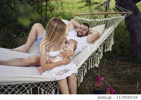 mother is breastfeeding her baby daughter in a hammock, father is lying next to her mother is breastfeeding her baby daughter in a hammock, father is lying next to her 117050538
