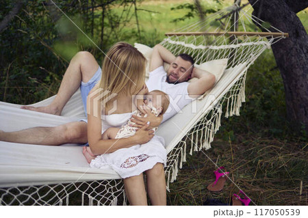 mother is breastfeeding her baby daughter in a hammock, father is lying next to her 117050539
