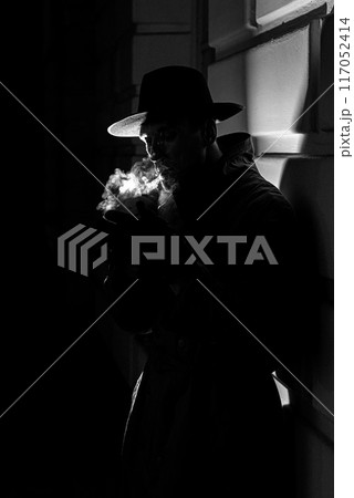 dark dramatic silhouette of a man in a hat Smoking a cigarette on the street at night dark dramatic silhouette of a man in a hat Smoking a cigarette on the street at night 117052414