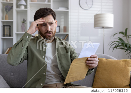 Worried man reading letter on couch at home, holding envelope, showing signs of stress and anxiety. Concerned expression indicates financial issues or bad news. 117056336