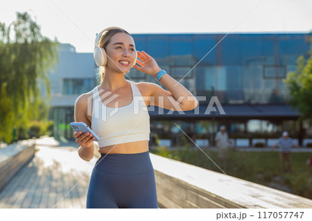 Happy young woman jogger listening to music via headphones and smartphone walking in city park 117057747