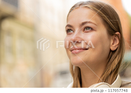 Thoughtful young woman tourist with wireless headphones around neck looking up and smiling outdoors 117057748
