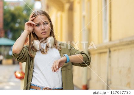 Serious dissatisfied young Caucasian woman standing and looking for someone waiting on city street 117057750
