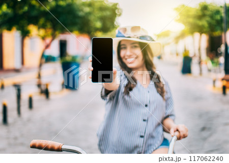 Smiling tourist on bicycle showing cell phone screen on the street. Beautiful girl with hat on bicycle showing cell phone screen on the street Smiling tourist on bicycle showing cell phone screen on the street. Beautiful girl with hat on bicycle showing cell phone screen on the street 117062240