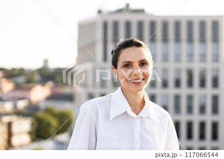 Smiling woman in white shirt standing outdoors in urban setting Smiling woman in white shirt standing outdoors in urban setting 117066544