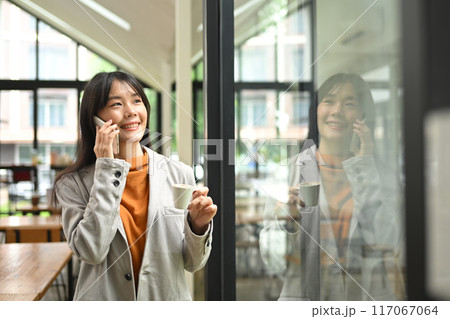 Charming young businesswoman having pleasant phone conversation while standing near office window 117067064