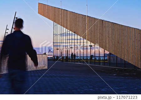 Blurred figure walks past modern architectural building in Tallinn Cruise Harbour with reflective glass at sunset. The evening sky and nearby structures are beautifully mirrored on the glass surface Blurred figure walks past modern architectural building in Tallinn Cruise Harbour with reflective glass at sunset. The evening sky and nearby structures are beautifully mirrored on the glass surface 117071223