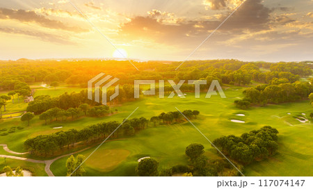 Golf course at sunset with beautiful sky and sand trap. Scenic panoramic view of golf fairway with bunker. Golf field with pines Golf course at sunset with beautiful sky and sand trap. Scenic panoramic view of golf fairway with bunker. Golf field with pines 117074147