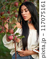 Gorgeous brunette female with long hair, picking apples in apple orchard. Portrait of Caucasian beauty wearing white chemise posing in garden with closed eyes in evening. Femininity, harvest concept. 117075161