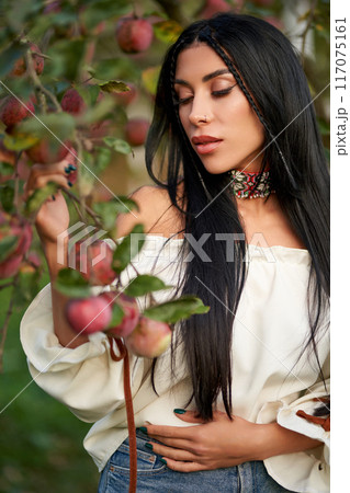 Gorgeous brunette female with long hair, picking apples in apple orchard. Portrait of Caucasian beauty wearing white chemise posing in garden with closed eyes in evening. Femininity, harvest concept. 117075161