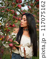Pensive brunette with bright necklace, picking apples in orchard in fall. Portrait of Caucasian beauty wearing white chemise posing in garden with closed eyes at day. Concept of femininity, harvest. 117075162
