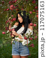 Thoughtful, serious woman in jeans shorts, touching apple tree branches, laden with fruits. Front view of young woman with long dark hair posing in apple orchard with closed eyes. Harvesting concept. 117075163