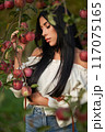 Serious young woman thinking, while leaning on apple tree branches at warm autumn day. Front view of calm Caucasian beauty with dark hair in apple orchard. Concept of emotions, female beauty, fall. 117075165