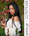 Attractive female with long dark hair, leaning on apple branch in orchard. Portrait of pensive beauty wearing stylish blouse posing in garden with closed eyes, in fall. Concept of beauty, harvesting. 117075166