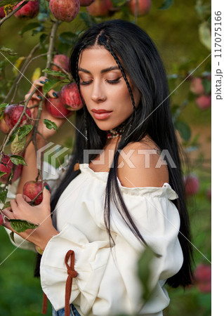 Attractive female with long dark hair, leaning on apple branch in orchard. Portrait of pensive beauty wearing stylish blouse posing in garden with closed eyes, in fall. Concept of beauty, harvesting. Attractive female with long dark hair, leaning on apple branch in orchard. Portrait of pensive beauty wearing stylish blouse posing in garden with closed eyes, in fall. Concept of beauty, harvesting. 117075166