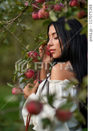Gorgeous relaxed woman leaning on apple tree branch in apple orchard. View from shoulder of tattooed beauty wearing white shoulderless blouse in garden at twilight. Concept of female beauty, harvest. Gorgeous relaxed woman leaning on apple tree branch in apple orchard. View from shoulder of tattooed beauty wearing white shoulderless blouse in garden at twilight. Concept of female beauty, harvest. 117075168