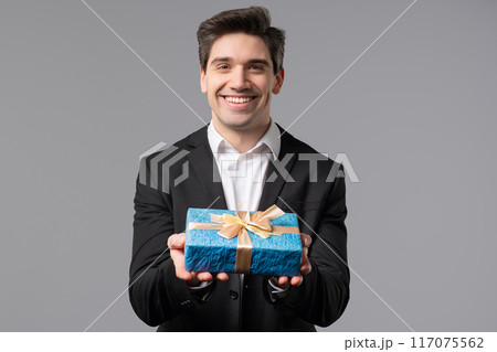 Handsome Businessman In Suit Gives Gift Box Present To Camera. Man is happy, smiling on grey office wall background. Positive mood, holiday, bonus, birthday concept. 117075562
