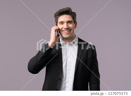 Adult 30s businessman speaks with smile on mobile phone. Guy holding and using smartphone. Formal suit outfit. Grey studio background 117075573