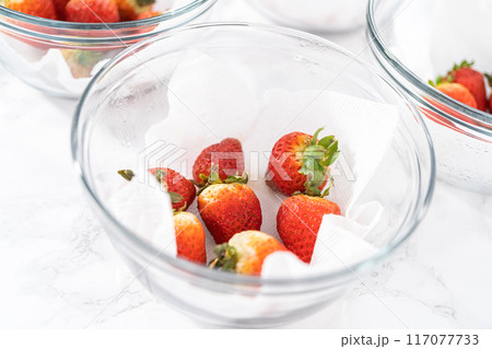 Bright red strawberries, interspersed with signs of mold, rest in a glass bowl lined with a paper towel on a white napkin, indicating improper storage techniques. 117077733