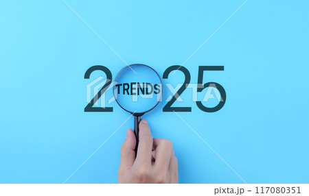 Business Trends 2025 Concept. A hand holds a magnifying glass focusing on the word "Trends" within the year "2025," representing the concept of future business and marketing trends and planning. 117080351