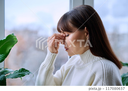 Middle aged woman experiencing headache migraine, at home near window Middle aged woman experiencing headache migraine, at home near window 117080637