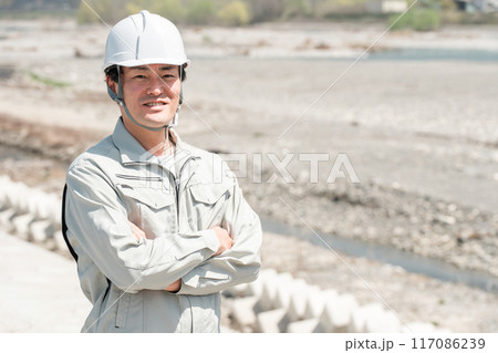 土木業・河川工事・建設現場に立つヘルメットを被った作業着姿のアジア人男性作業員 土木業・河川工事・建設現場に立つヘルメットを被った作業着姿のアジア人男性作業員 117086239