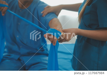 Close up hand patient doing stretching exercise with a flexible exercise band and a physical therapist hand to help in clinic room. 117087984