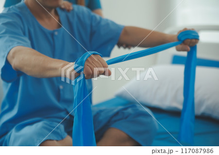 Close up hand patient doing stretching exercise with a flexible exercise band and a physical therapist hand to help in clinic room. Close up hand patient doing stretching exercise with a flexible exercise band and a physical therapist hand to help in clinic room. 117087986