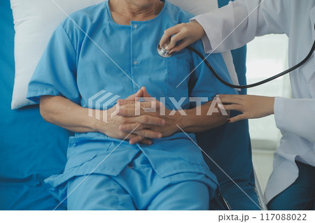 Doctor using stethoscope to listen senior man heart during examination in hospital room and wearing visor as safety precaution against coronavirus. Medical control for infections, disease. 117088022