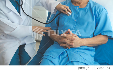 Doctor using stethoscope to listen senior man heart during examination in hospital room and wearing visor as safety precaution against coronavirus. Medical control for infections, disease. 117088023