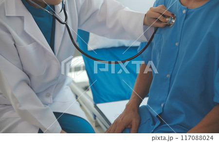 Doctor using stethoscope to listen senior man heart during examination in hospital room and wearing visor as safety precaution against coronavirus. Medical control for infections, disease. 117088024