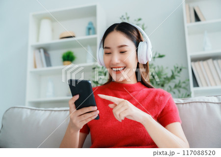 Young Asian woman relaxing on sofa at home, listening music with headset on cell phone 117088520